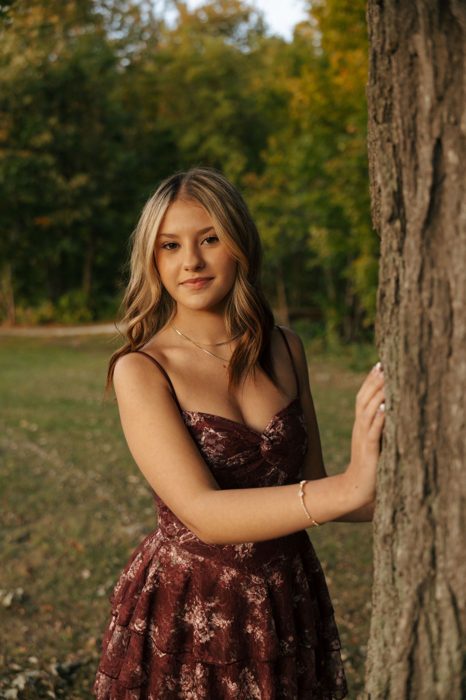 Young blonde woman in burgundy floral dress leaning against a tree in golden hour light.