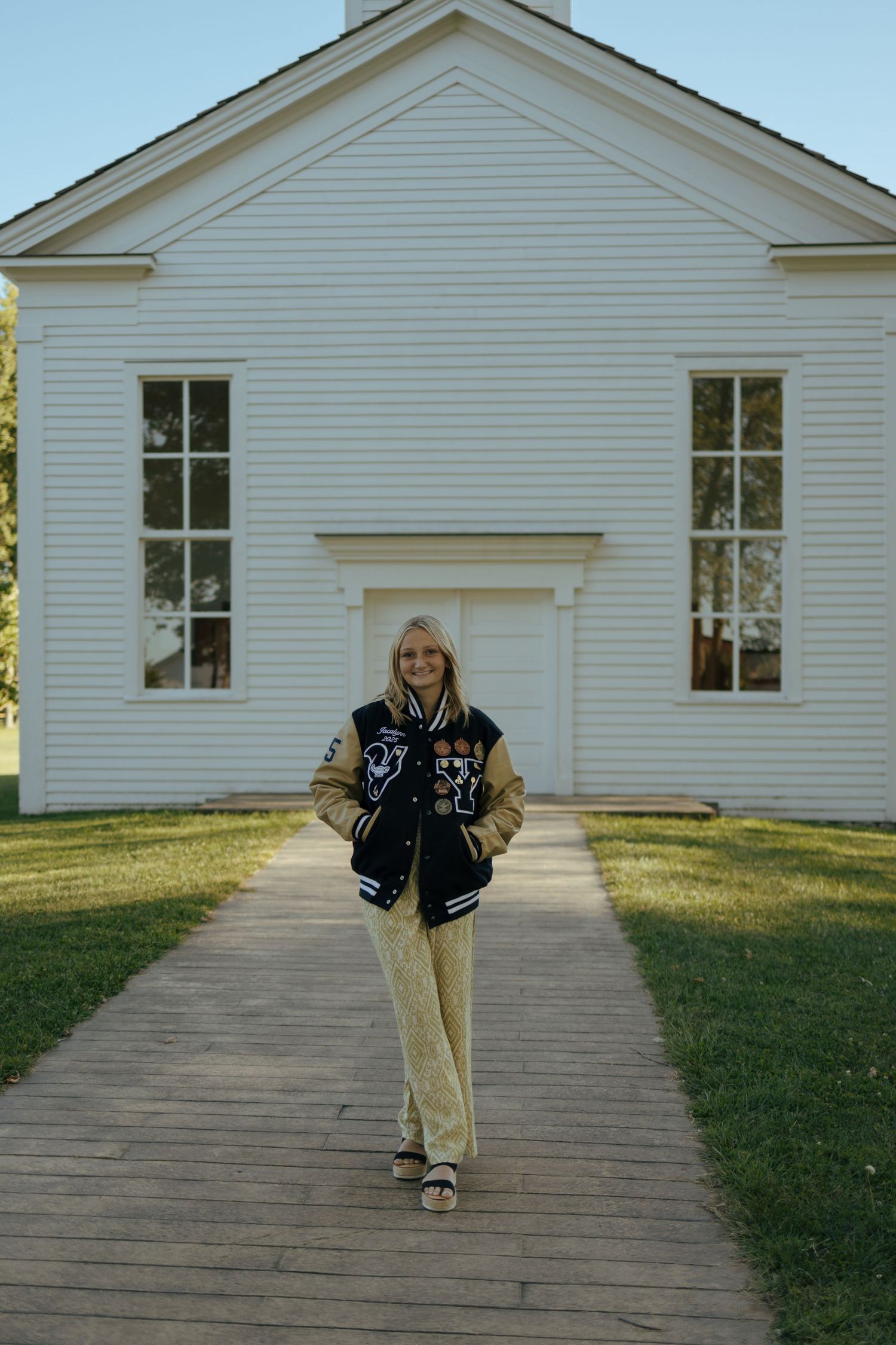 Blonde teen in varsity jacket and gold pants walking path toward white historic church building.