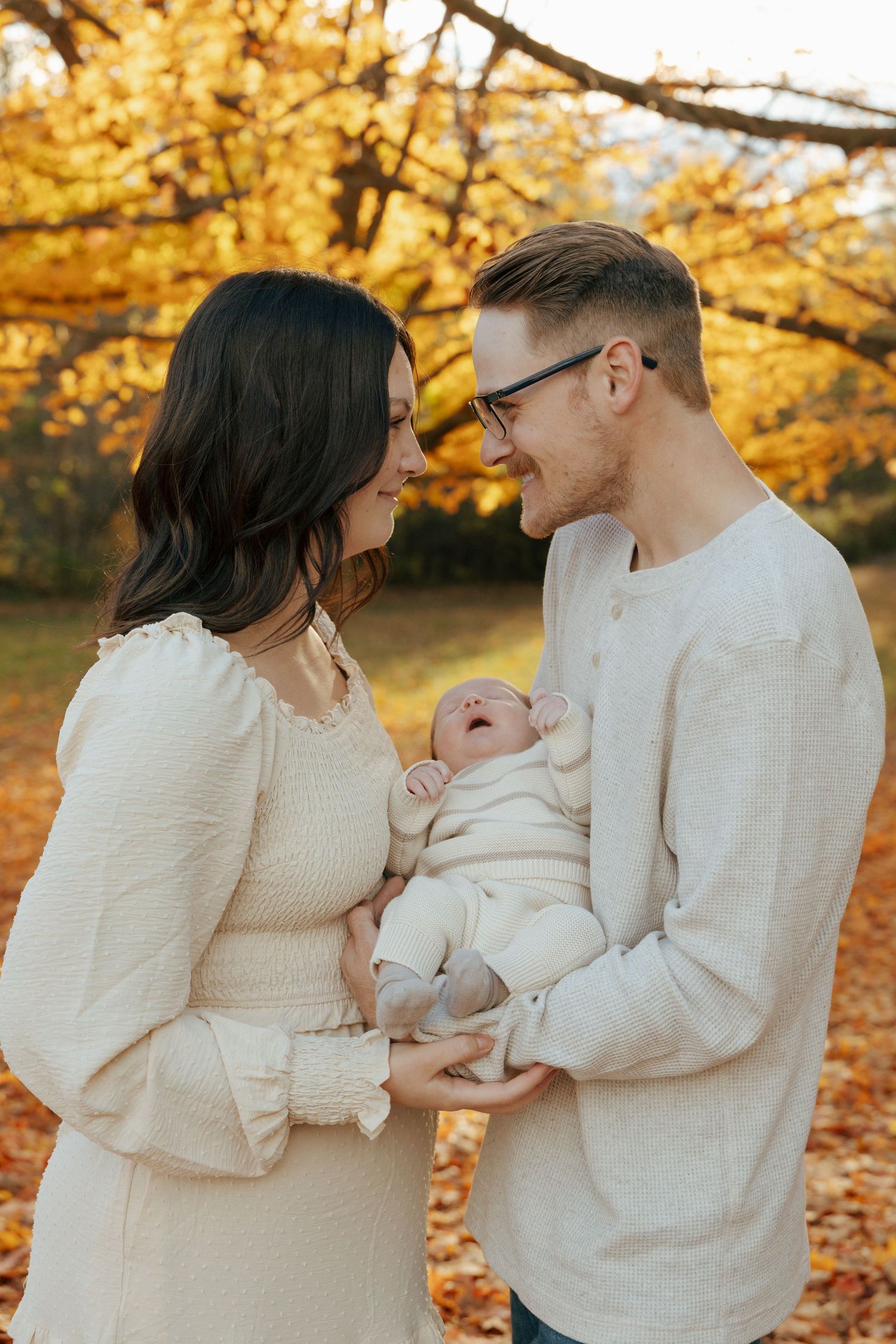 Young couple smiling at newborn baby they hold together under golden autumn foliage.