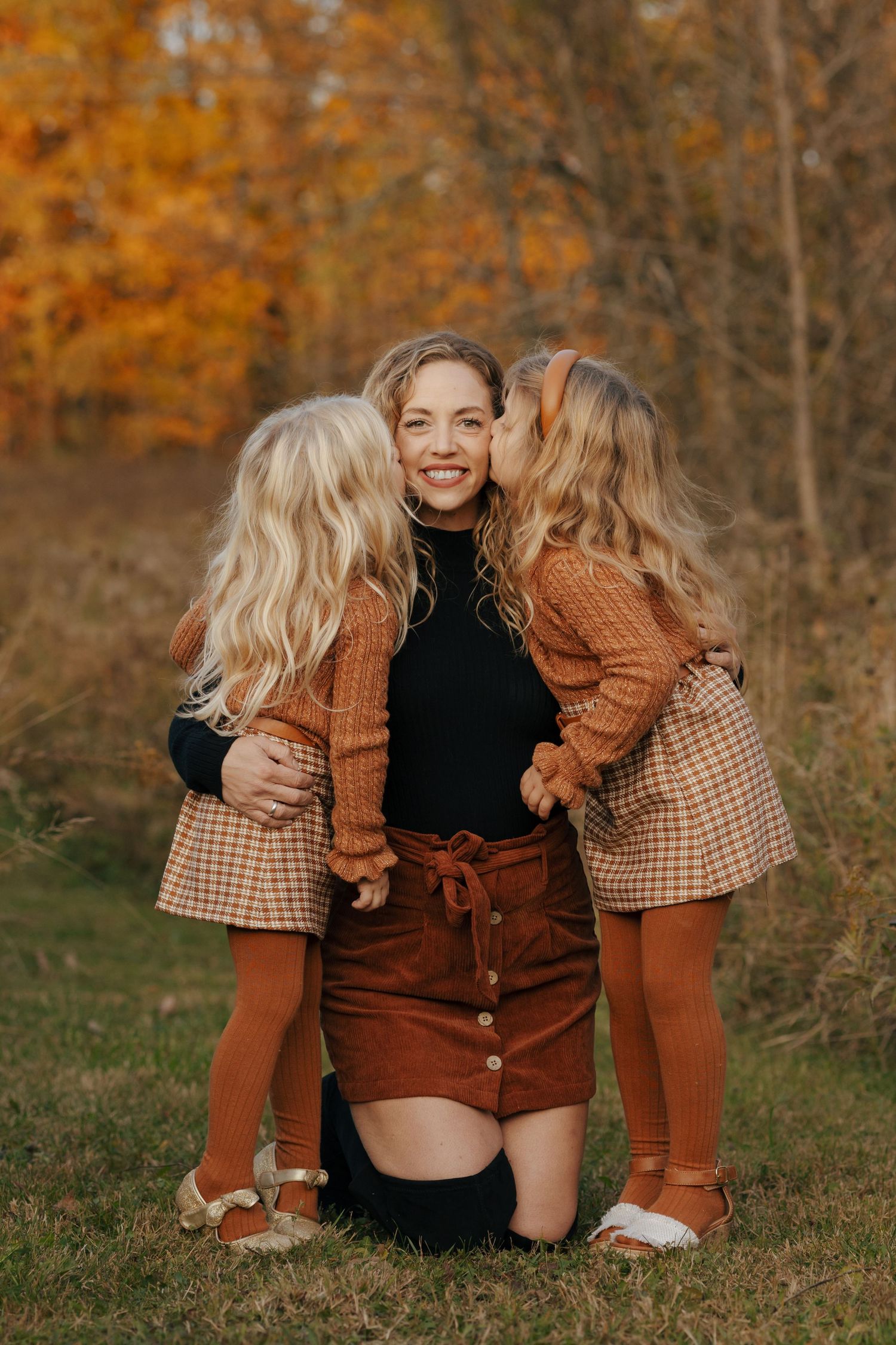 Mother kneeling outdoors with two blonde daughters in matching plaid outfits during autumn fall session.