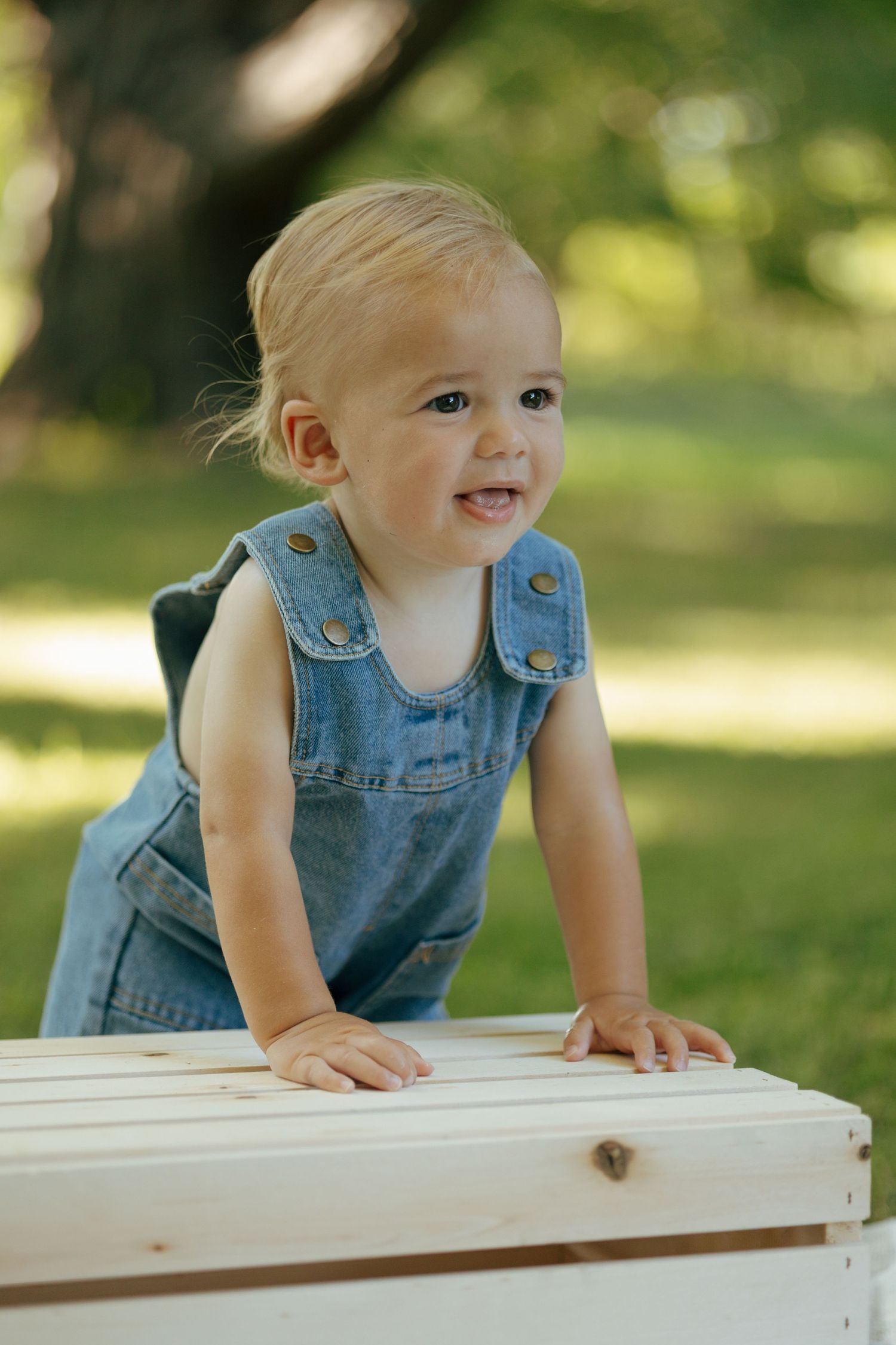 Smiling baby in denim overalls leaning on a white wooden box outdoors.