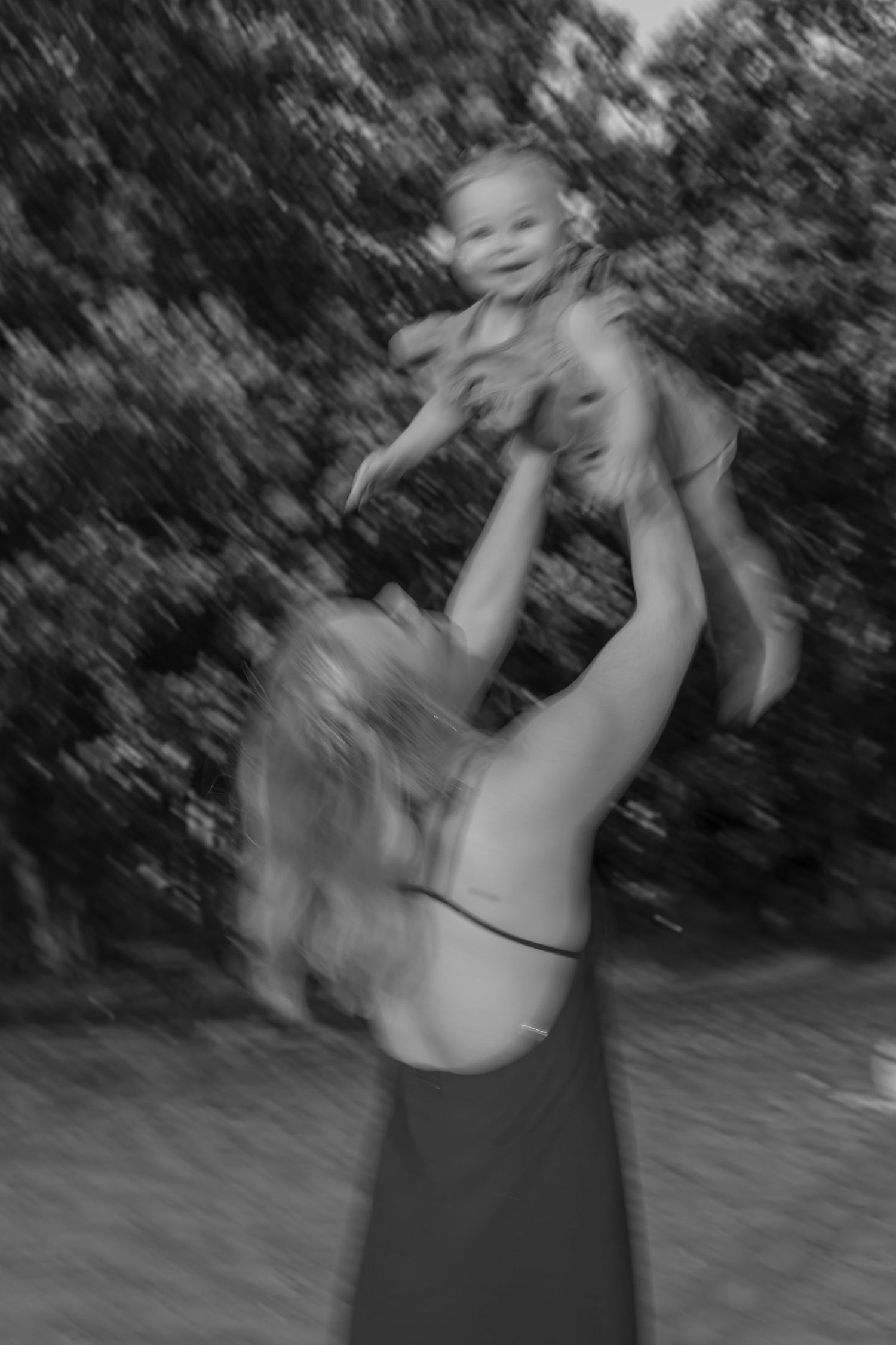 A joyful black and white photo of a mother lifting her laughing baby high into the air outdoors.