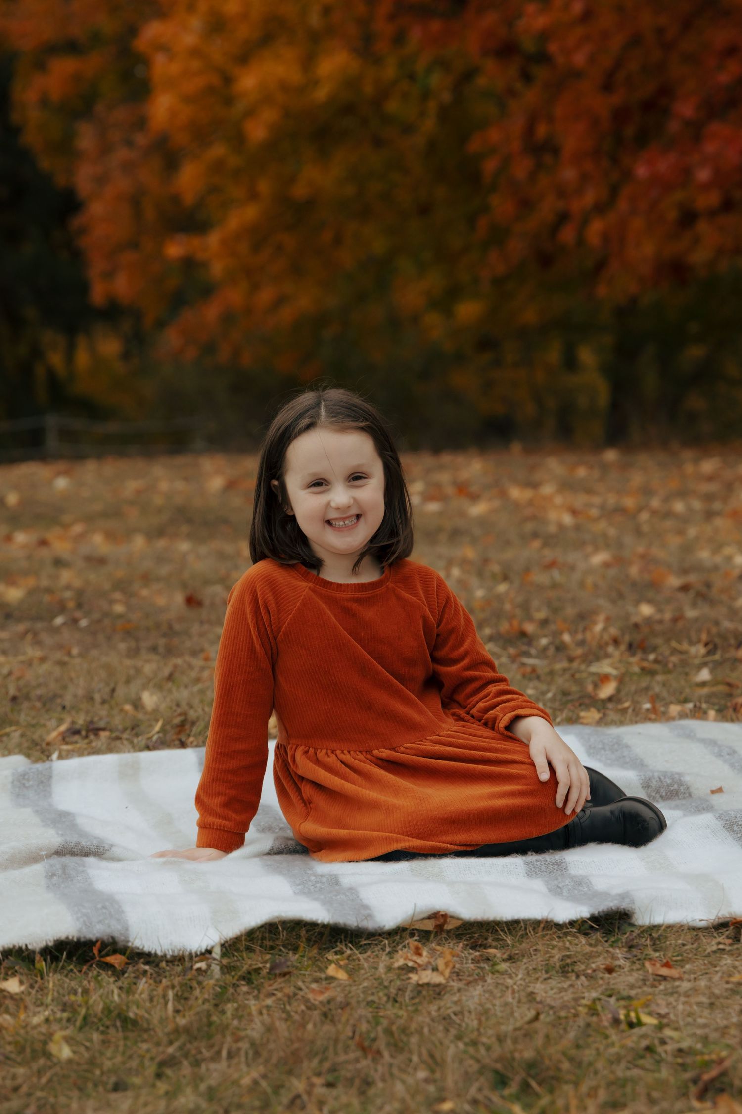 Young girl in rust orange dress sitting on plaid blanket with colorful fall trees behind her.