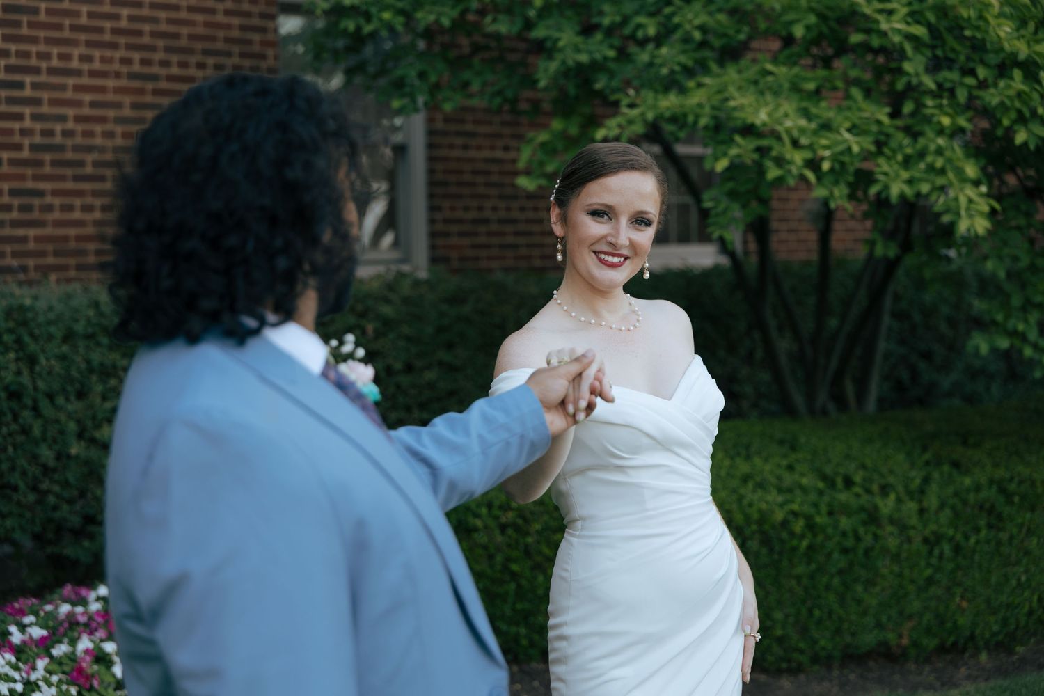 Smiling bride in white gown holding hands with groom in light blue suit in outdoor garden setting.