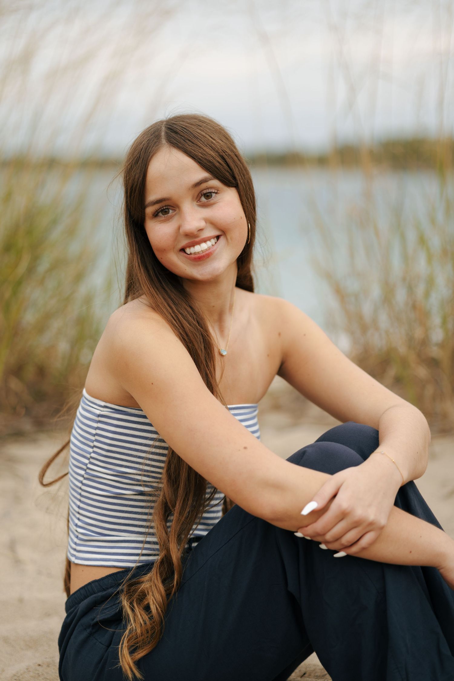 Smiling young woman in striped tube top sitting on sandy beach with tall grass and water behind her.