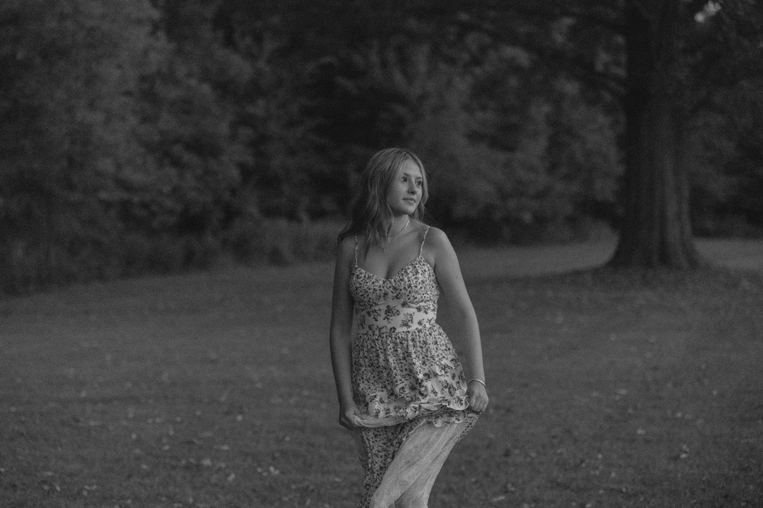 Black and white photo of a blonde woman in a floral dress walking in a park.