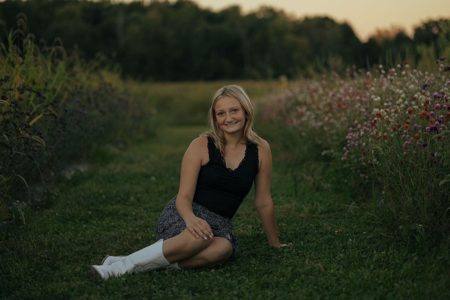 Smiling blonde teen girl in black tank and plaid skirt sitting on grass in wildflower field.
