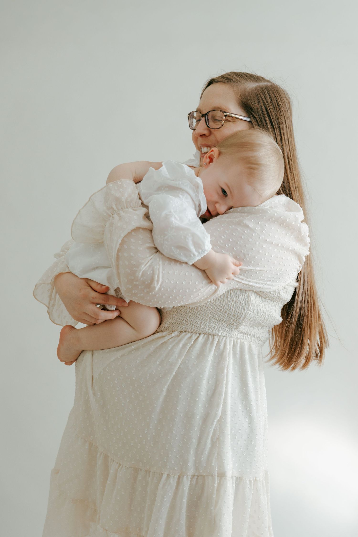 Joyful pregnant woman in white dress cuddling laughing baby during maternity photoshoot.