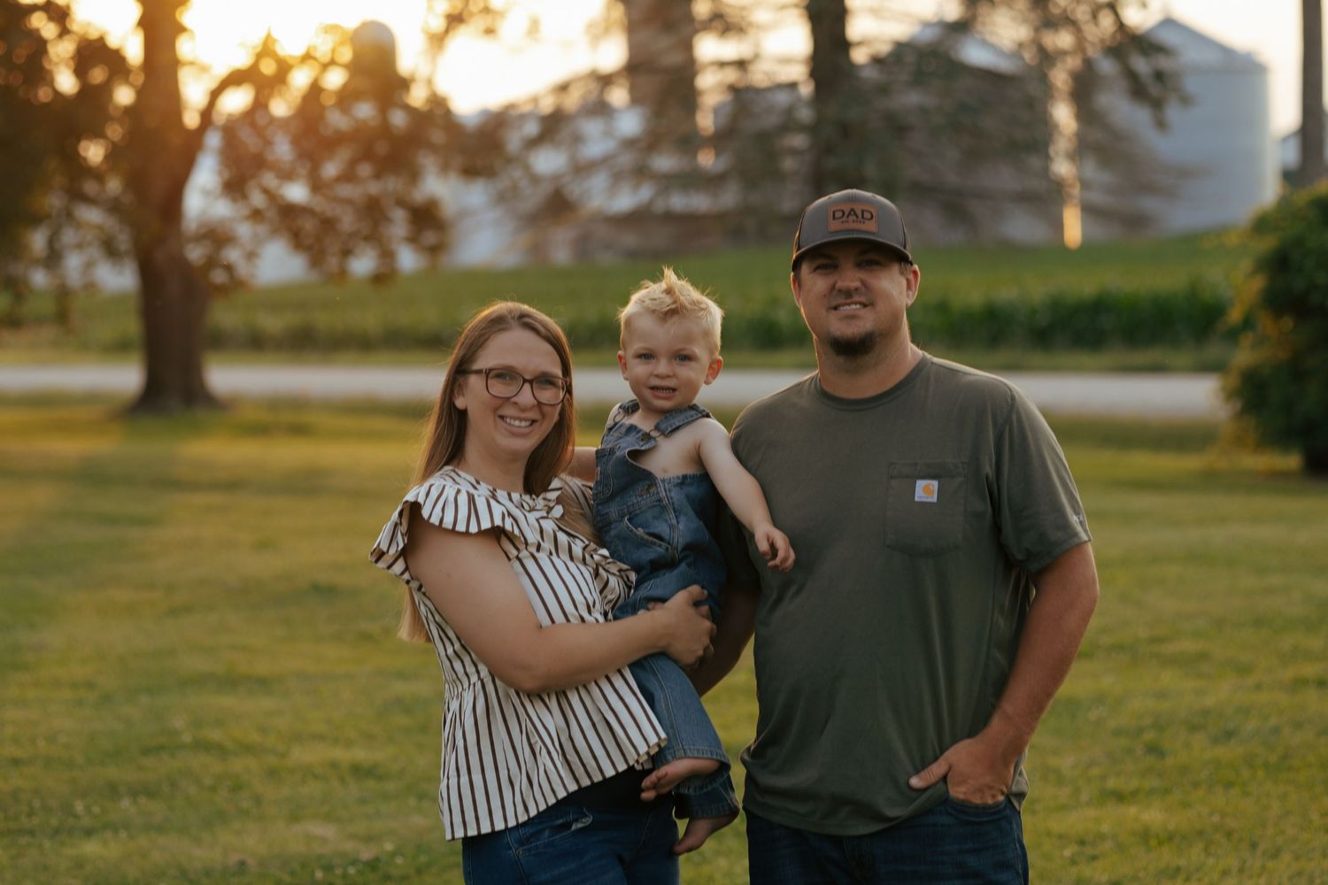 Young farm family posing at sunset with grain silos and green fields in the background.