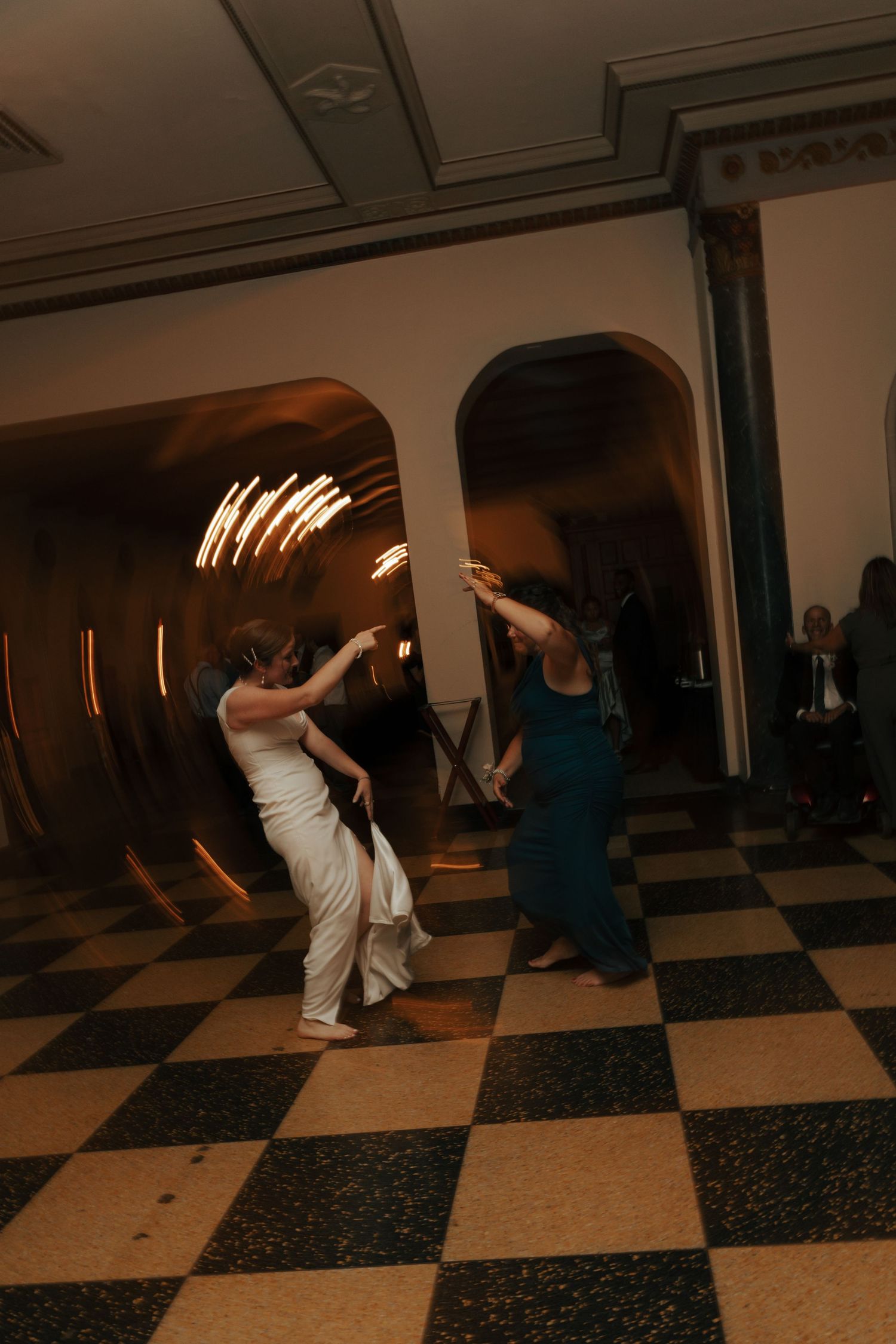 Bride and bridesmaid dancing joyfully in an elegant ballroom with arched doorways and checkered floors.