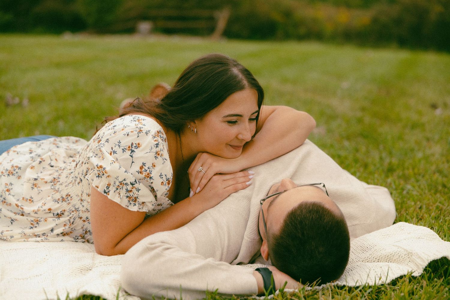Woman lying on blanket embracing man during romantic outdoor engagement photo session on grass.