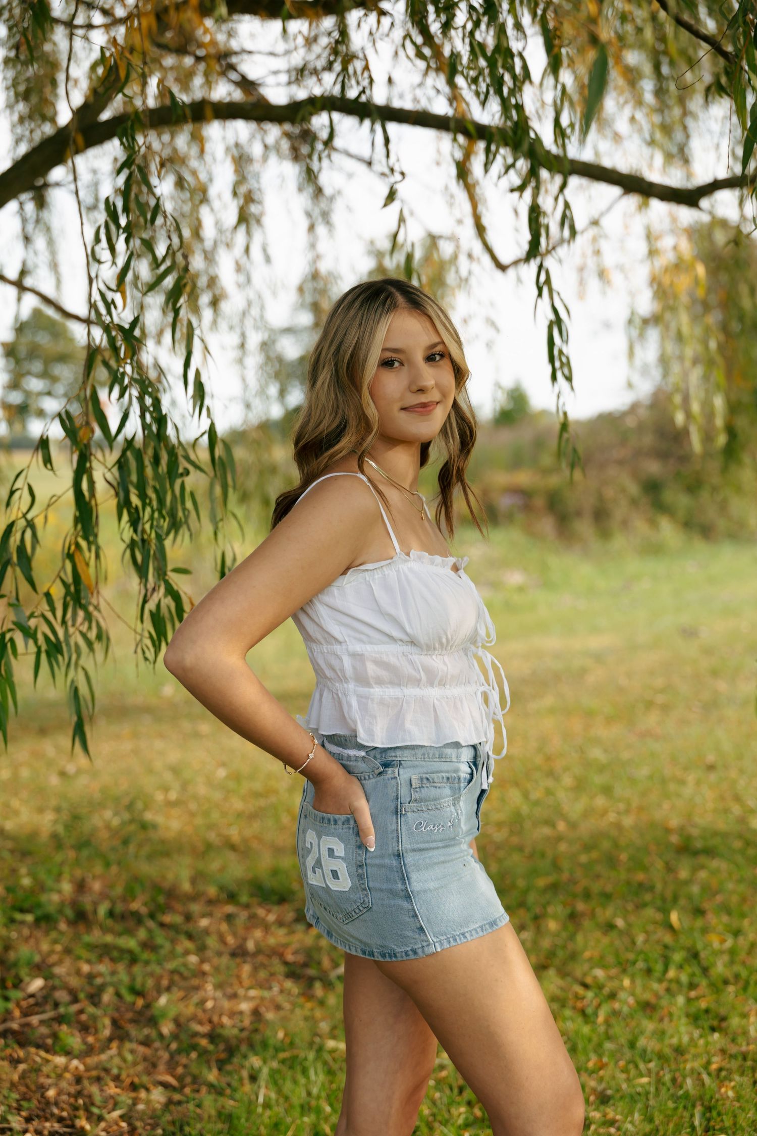 Teen girl in white ruched crop top and number 26 denim skirt posing under weeping willow in colorful fall park.