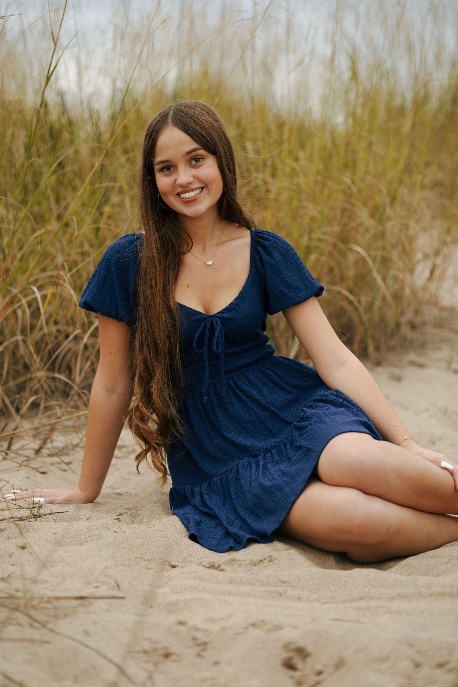 Young woman in navy blue dress sitting on sand smiling, surrounded by tall beach grass.