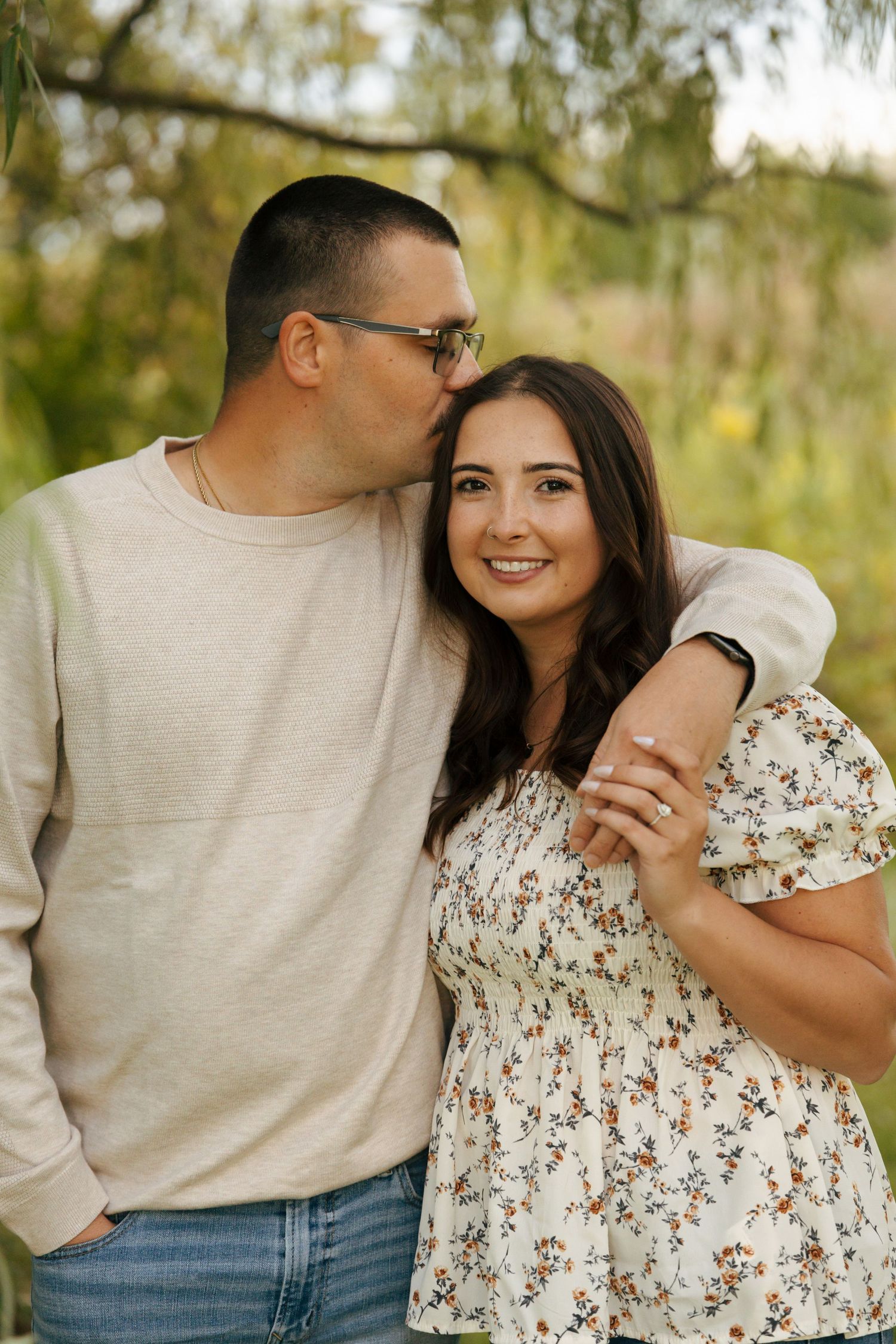 Couple poses outdoors, man kissing woman's temple while she smiles, wearing floral dress with engagement ring visible.