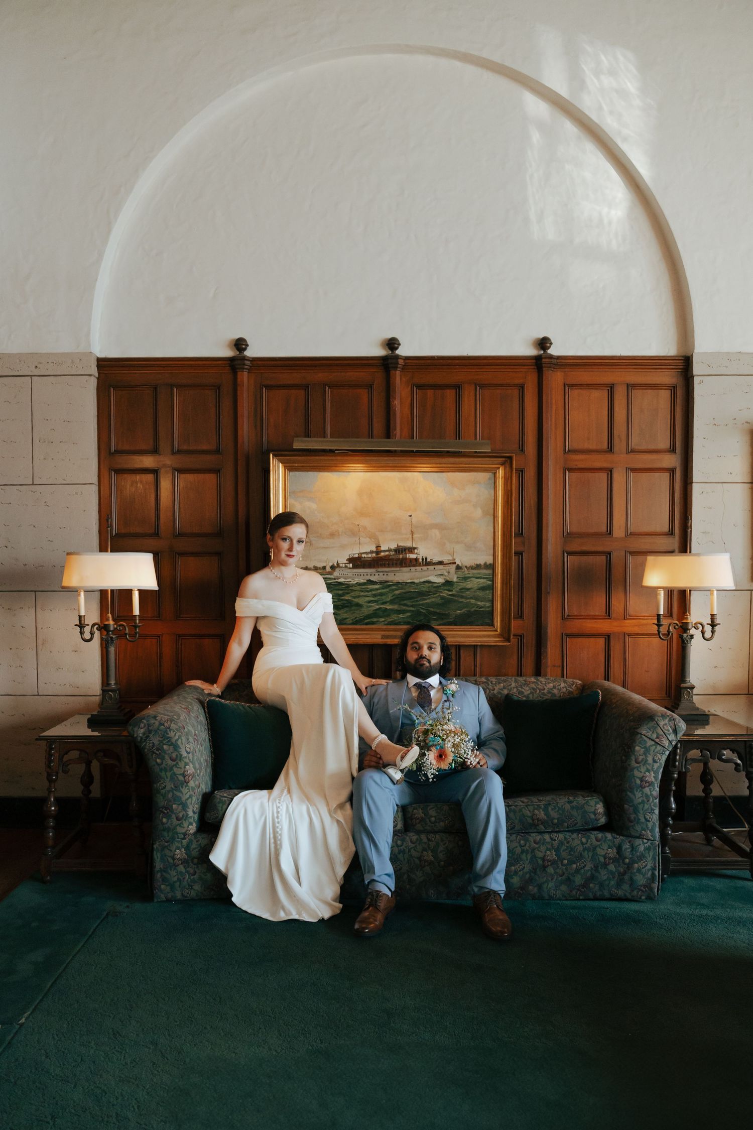 Bride and groom sitting on elegant sofa before wooden paneled wall with painting in classic interior.