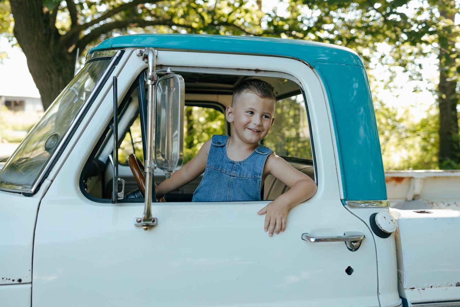 Happy young boy in denim overalls leans out window of a classic vintage pickup truck.