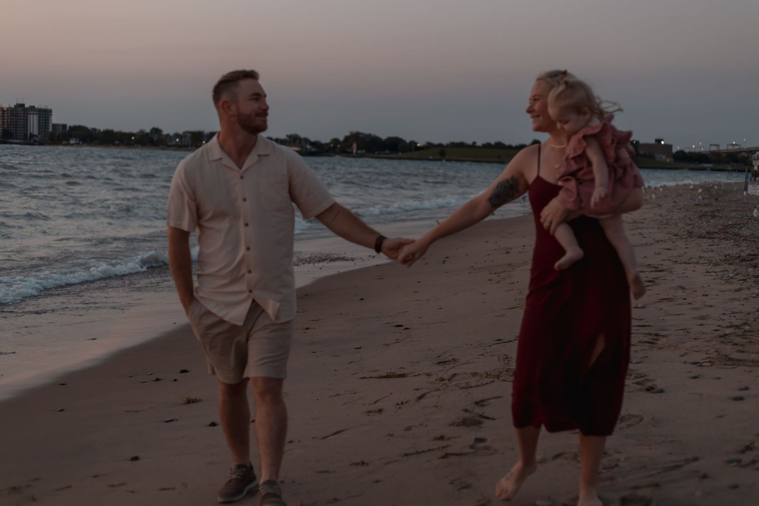 A happy couple holding hands and carrying their baby while walking along a sandy beach at dusk.