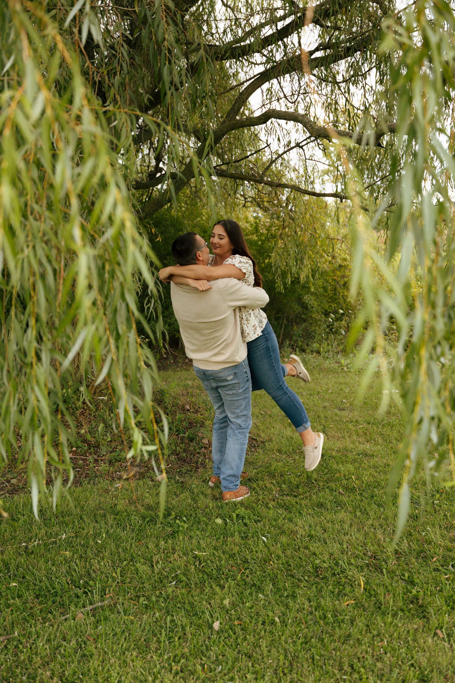 Couple embracing and spinning joyfully under a weeping willow tree in a green park.