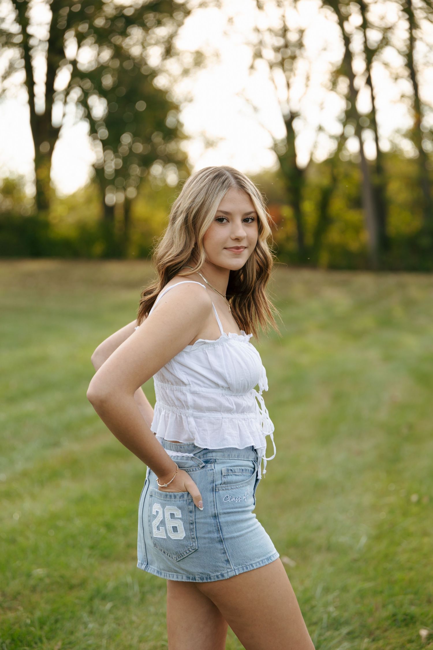 Blonde teen girl in white crop top and number 26 denim skirt posing confidently in golden sunlit open field.