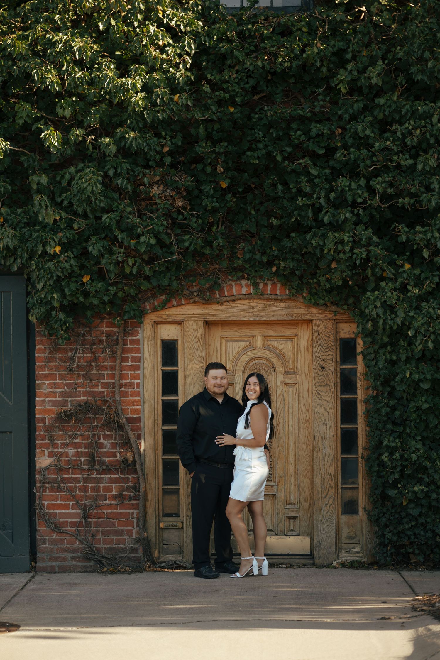 Smiling couple embracing in front of a wooden door on an ivy-covered brick building.