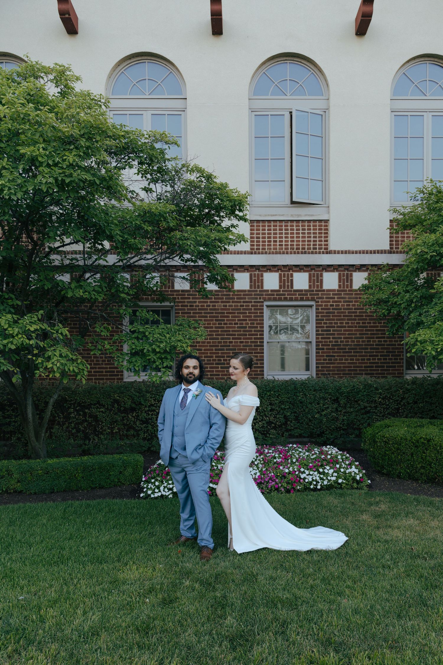 Bride in elegant white train gown and groom in blue suit standing on lush green lawn.