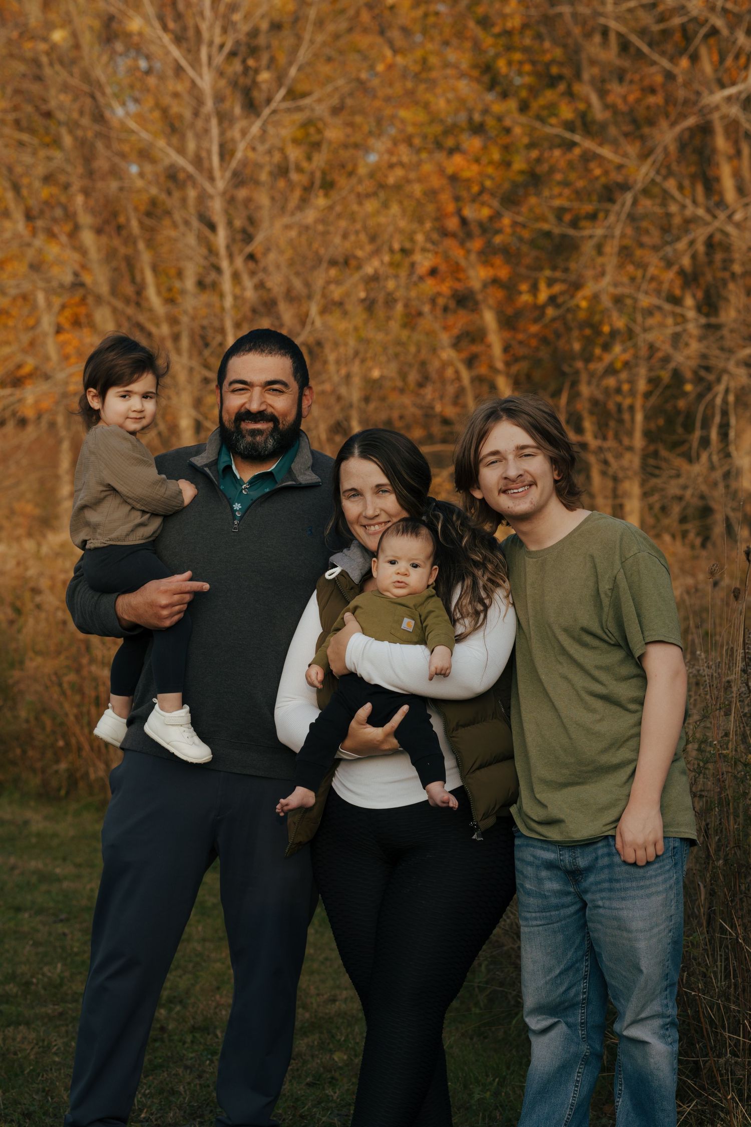 Family of five smiling together outdoors with colorful fall foliage background during autumn portrait session.
