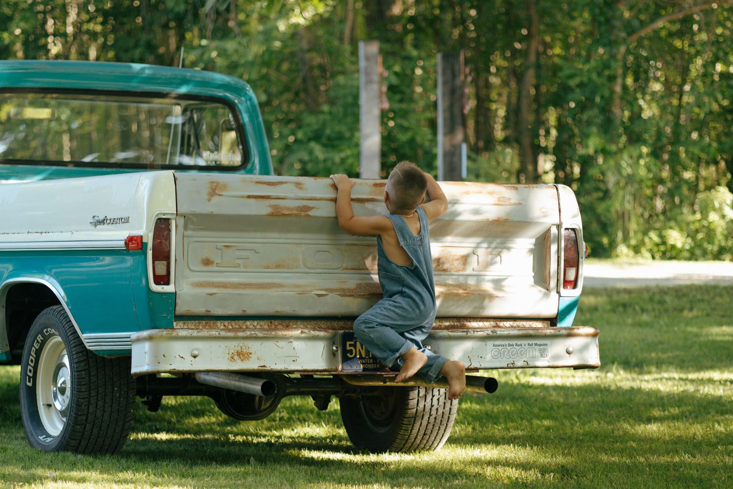 Young boy in overalls climbing into bed of vintage teal and white pickup truck.