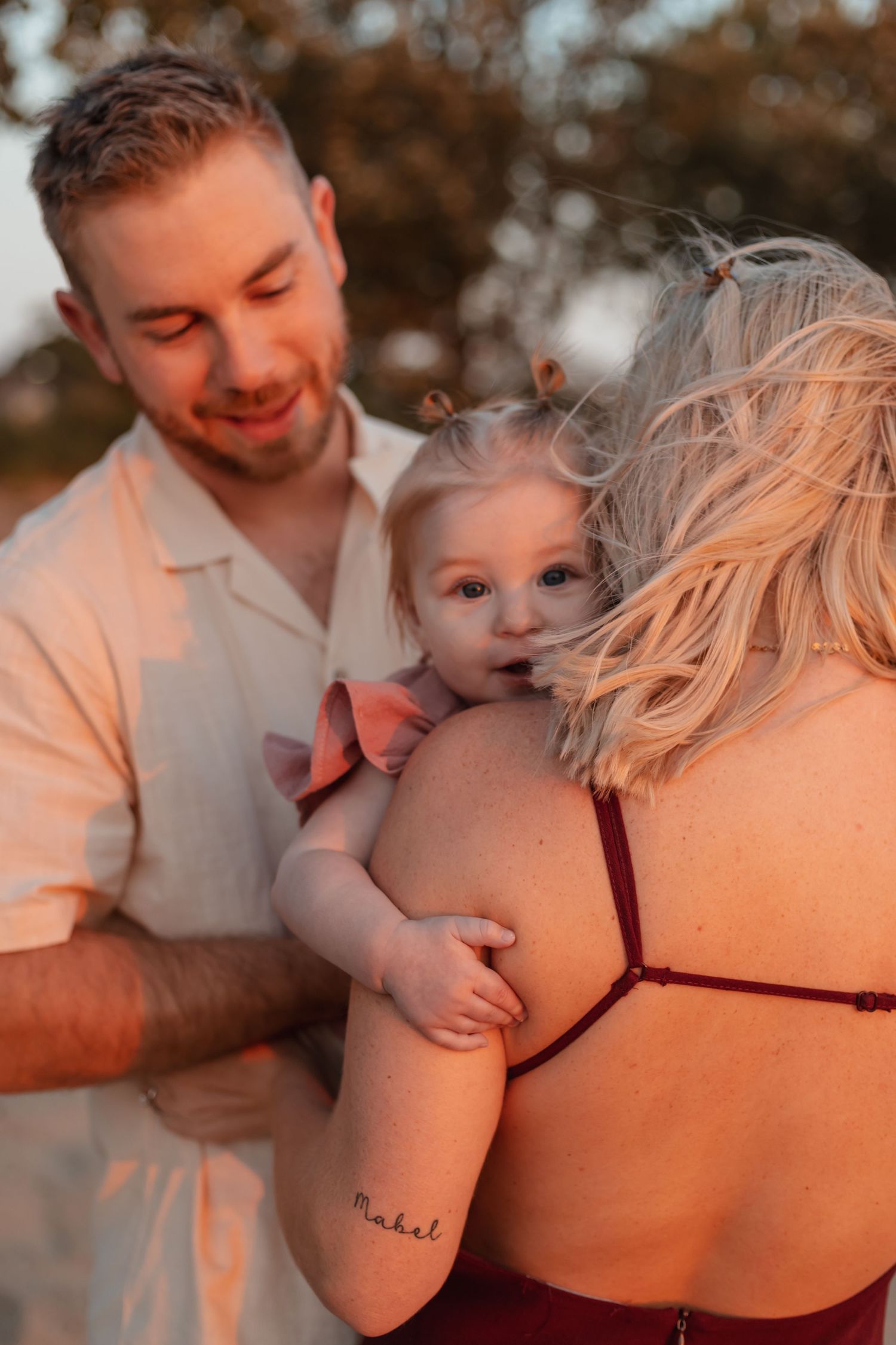 A father and mother lovingly hold their baby girl in warm golden sunset light outdoors.