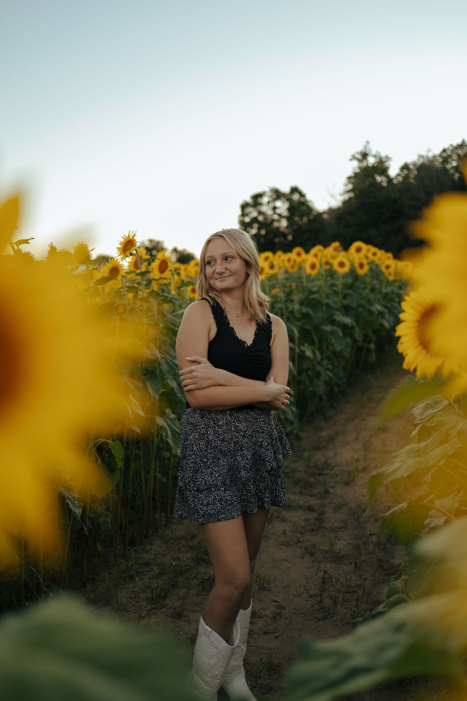 Blonde teen in black top, floral skirt and white cowboy boots standing in sunflower field at dusk.