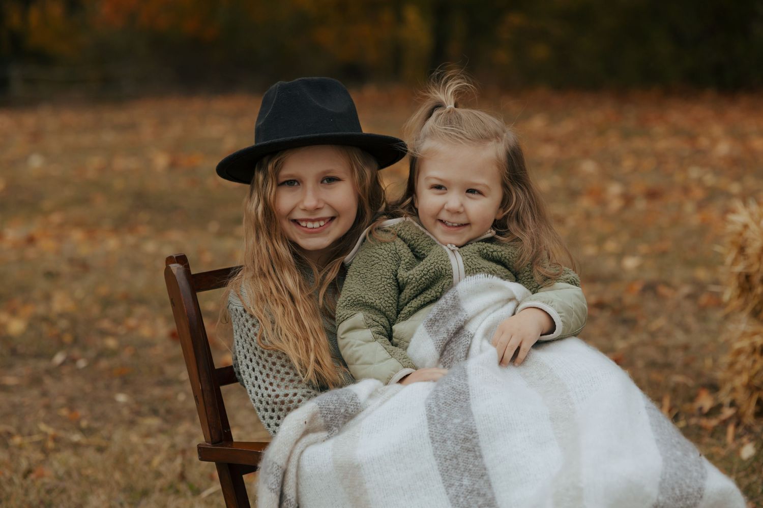 Two young sisters smiling together on wooden chair wrapped in blanket among autumn leaves.