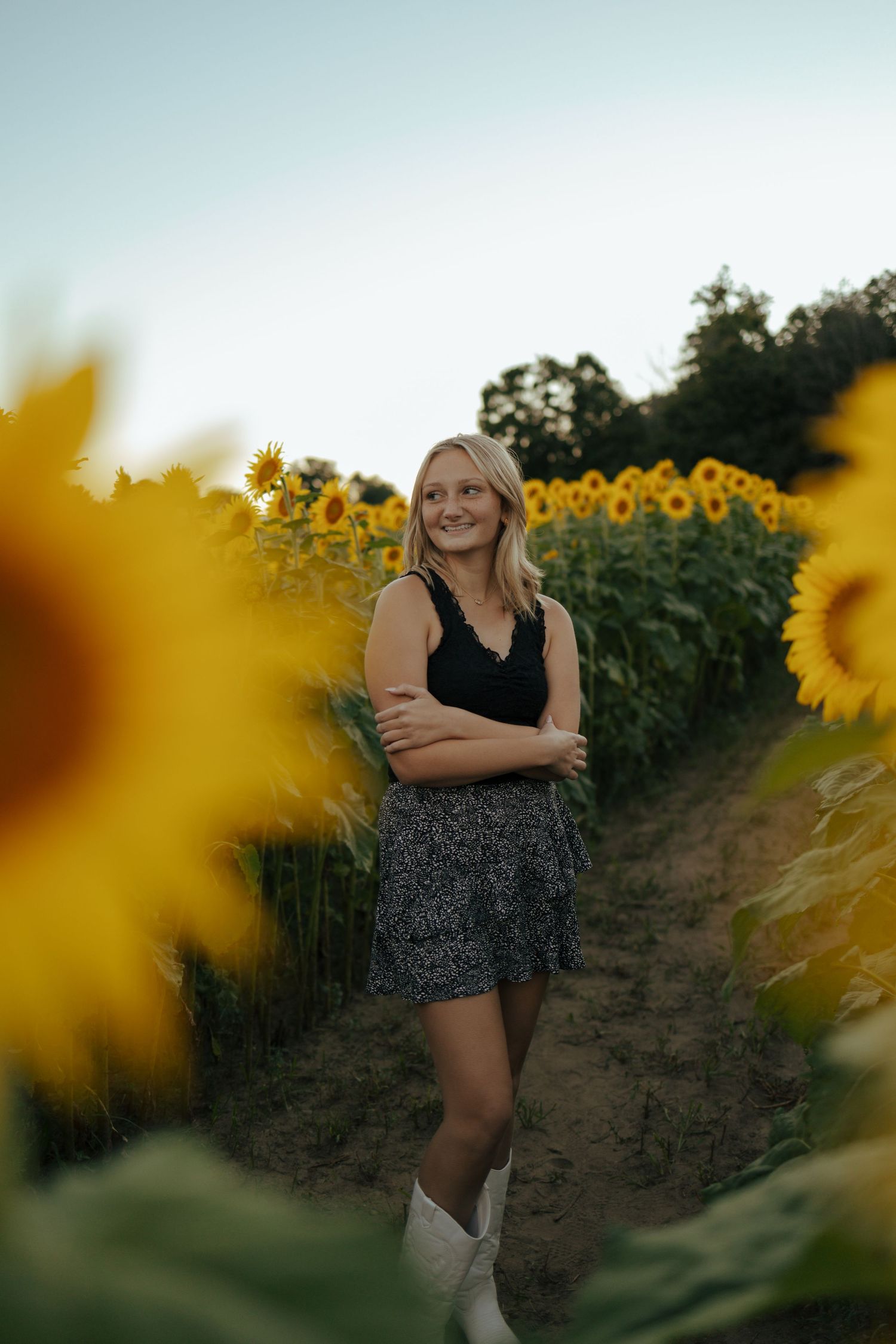 Smiling blonde woman in black top and floral skirt standing in a vibrant sunflower field at dusk.