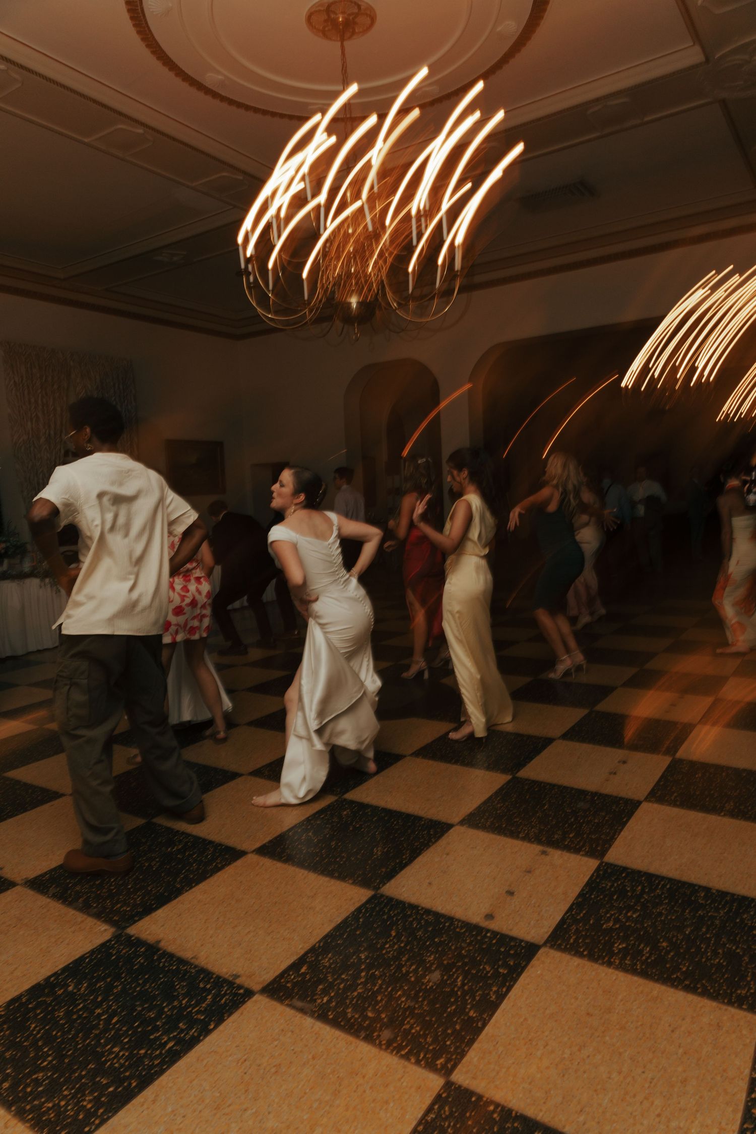 Bride and wedding guests dancing energetically on a black and white checkered floor under a chandelier.