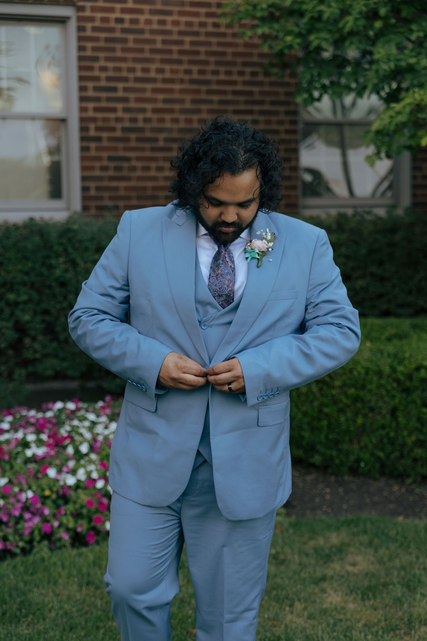 Groom in light blue suit adjusting jacket buttons while standing near colorful flower garden.