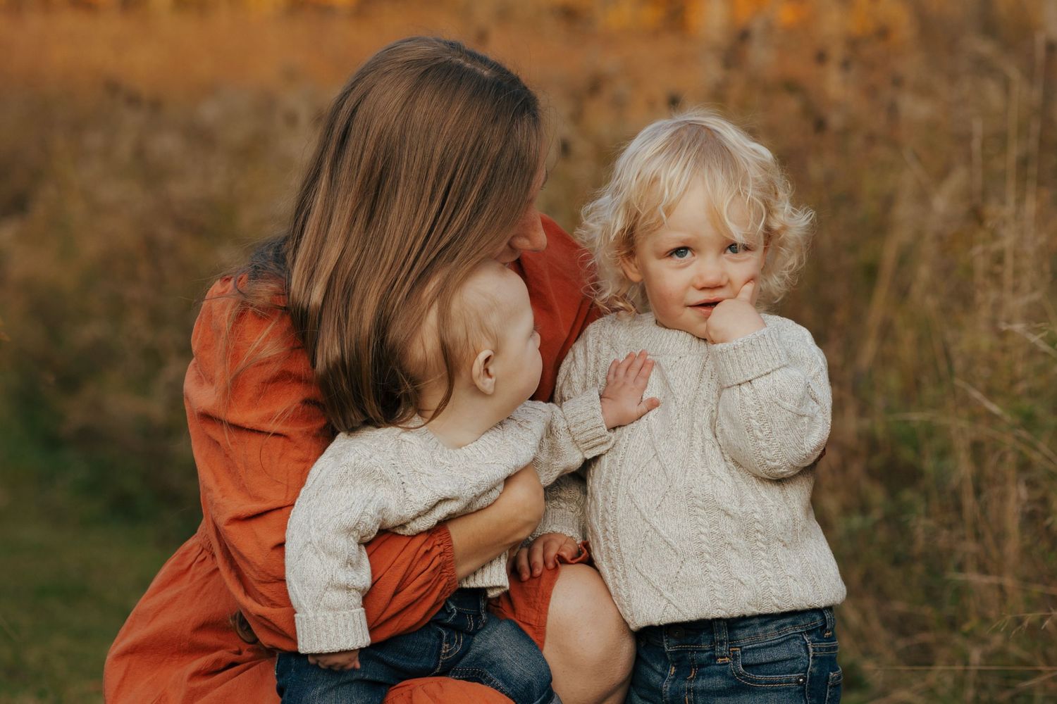 Mother in red dress tenderly holding two young toddlers in cream sweaters in autumn field.