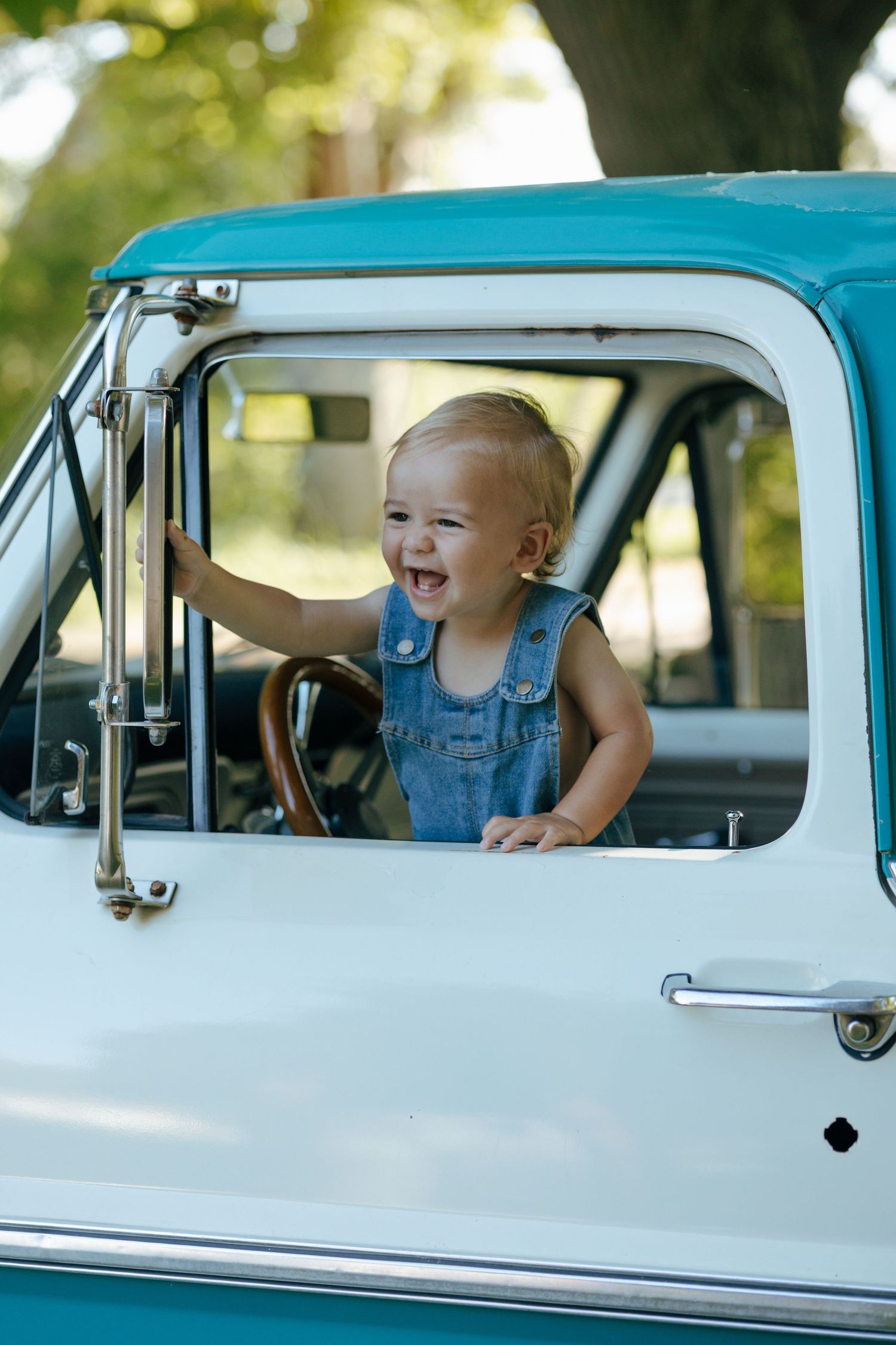 Happy toddler in denim overalls standing in driver seat of classic teal pickup truck.