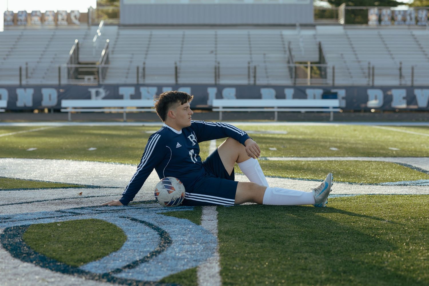 Soccer player #17 in navy Adidas uniform sits relaxed on turf field with ball at stadium.