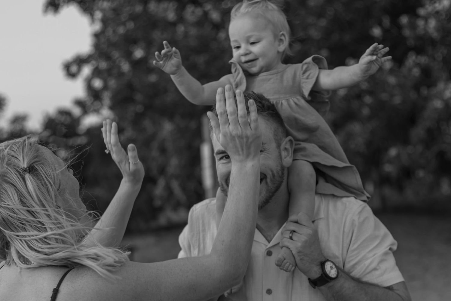 Black and white photo of parents lifting smiling baby girl on shoulders in park.
