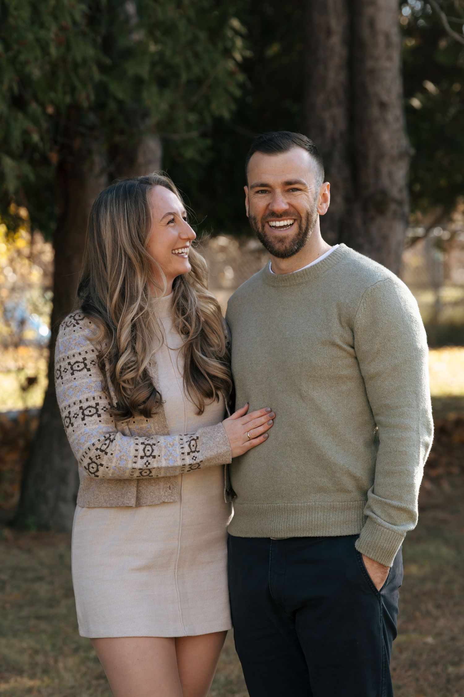 Happy couple laughing together outdoors among tall pine trees during fall engagement photos.