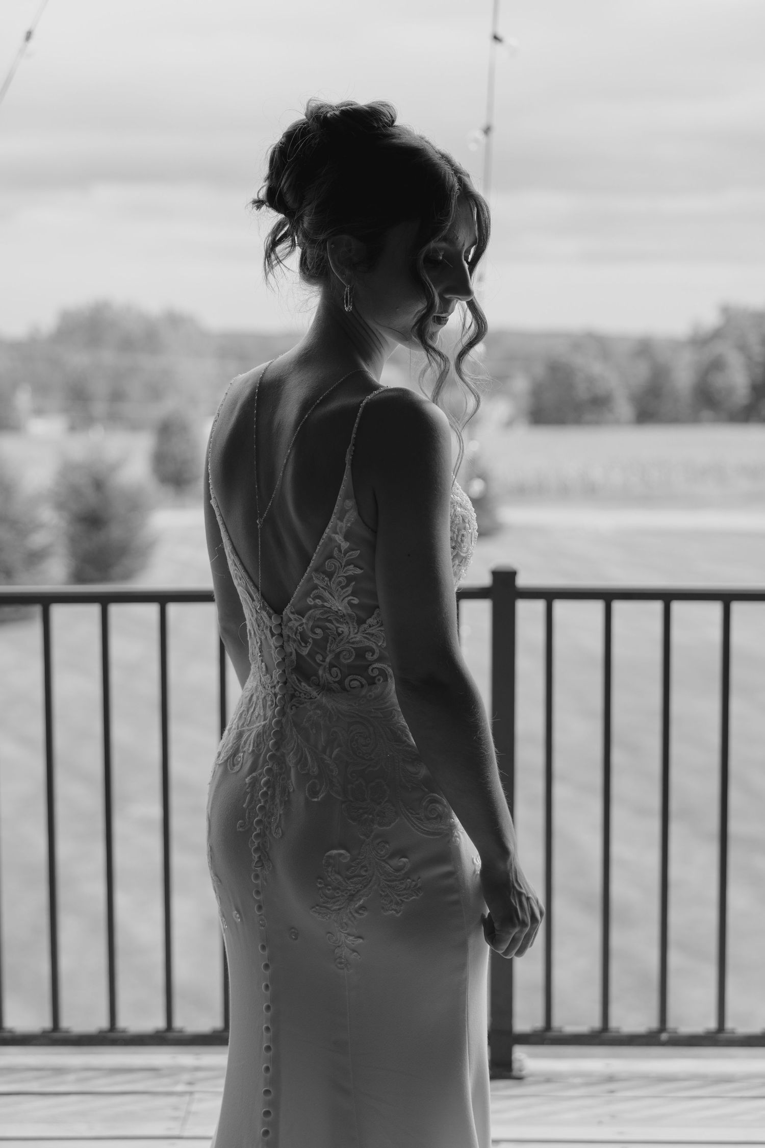 Elegant bride in a low back lace wedding gown stands on a balcony overlooking a scenic landscape.