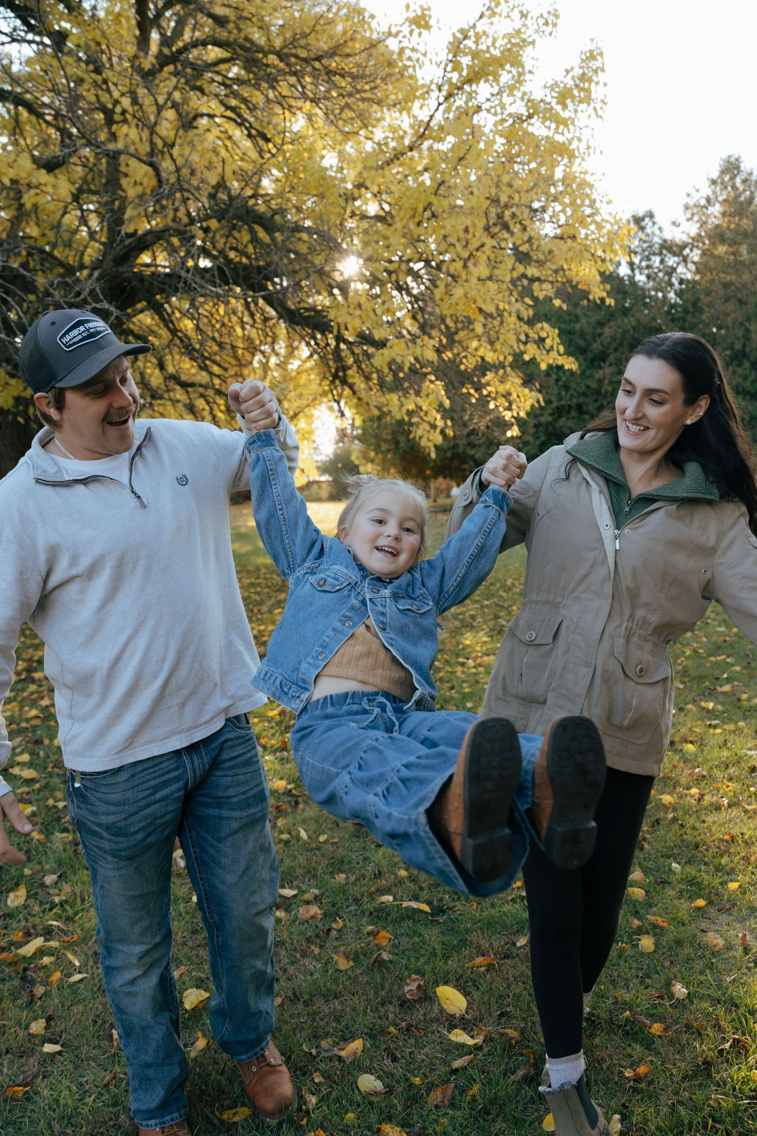Parents joyfully swinging their laughing daughter between them in a sunlit autumn park with yellow trees.