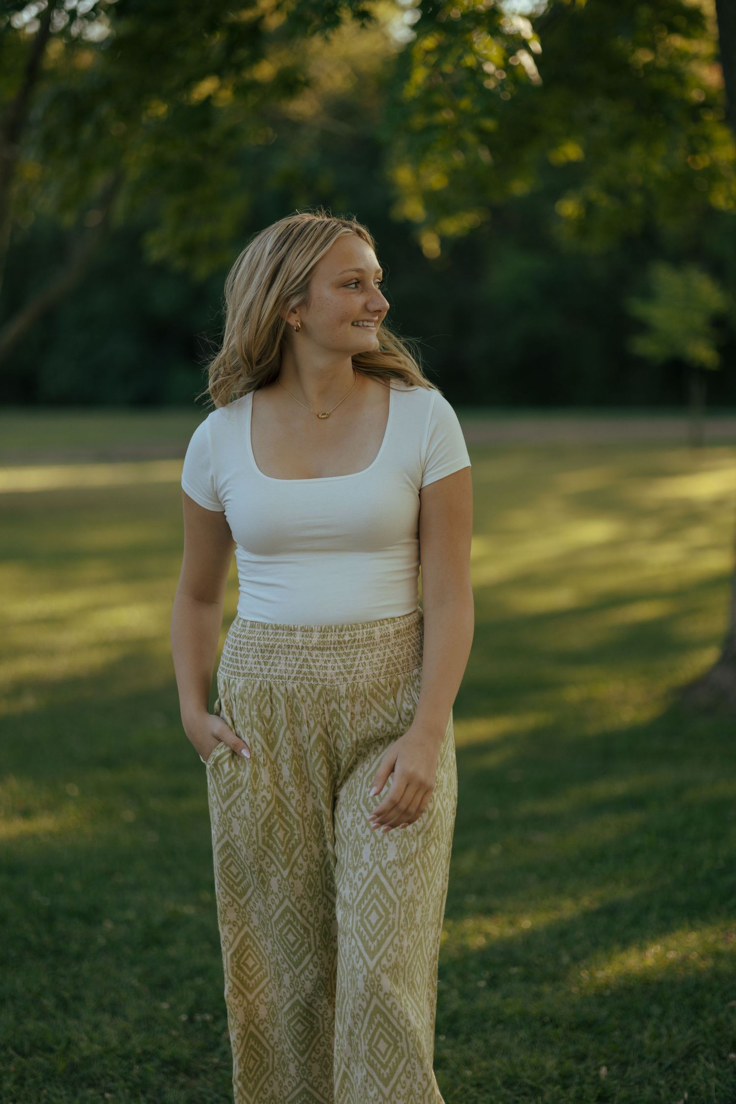 Blonde teenage girl in white top and gold patterned wide-leg pants standing in sunlit green park.