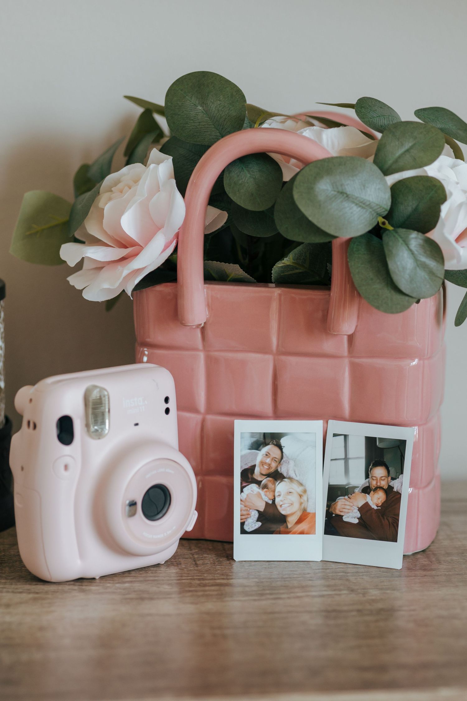 Pink Instax Mini camera beside polaroid photos and a pink ceramic basket with flowers and eucalyptus.