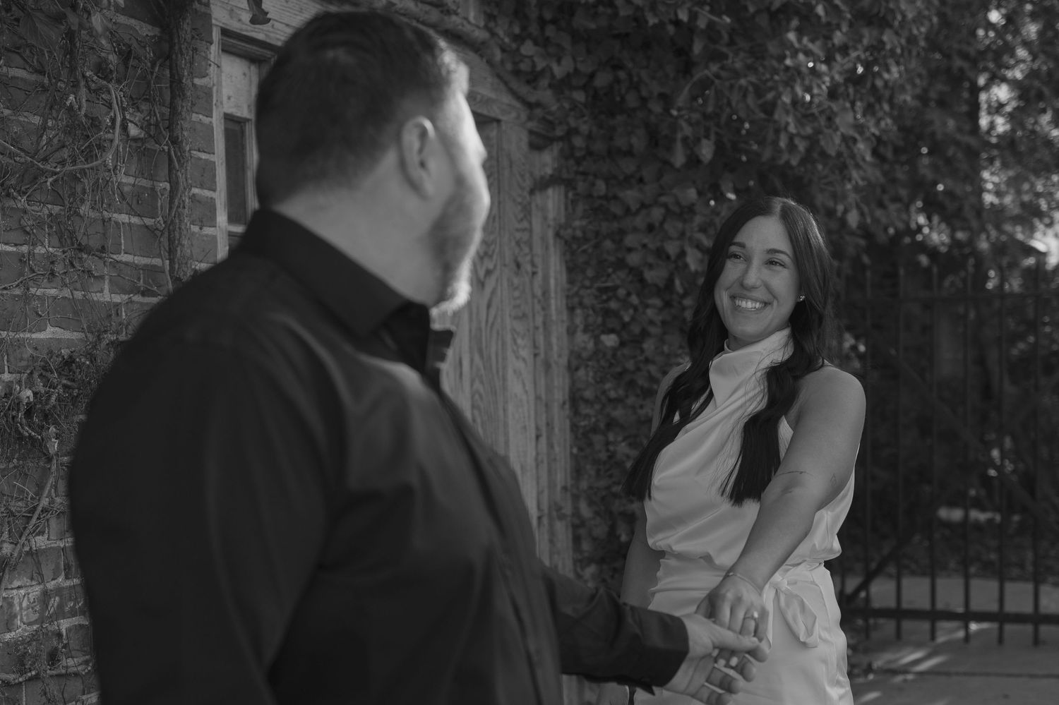 Black and white photo of a smiling couple holding hands near a brick wall with ivy.