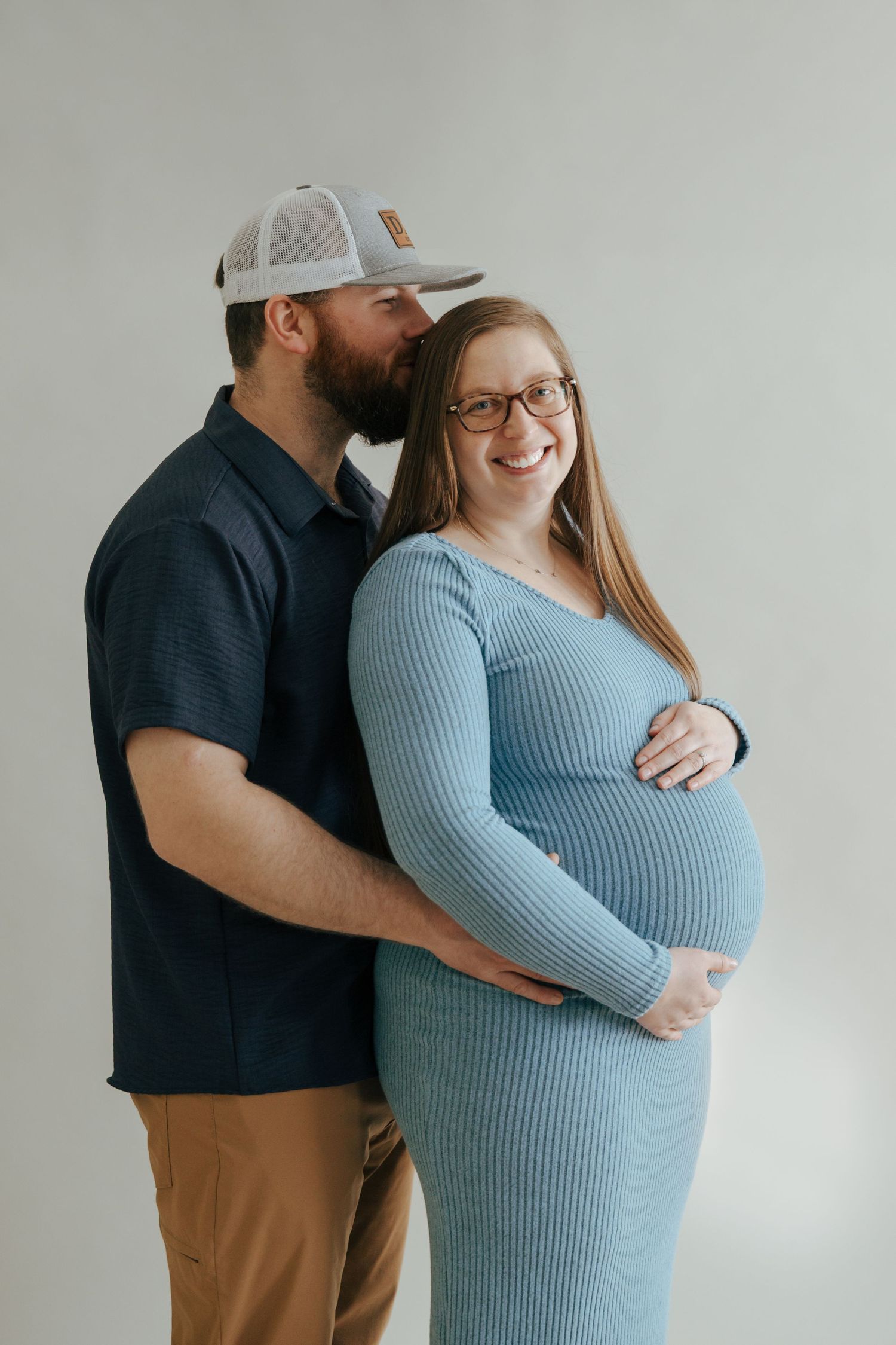 Pregnant couple posing indoors, man in navy shirt kissing woman in blue maternity dress on temple.