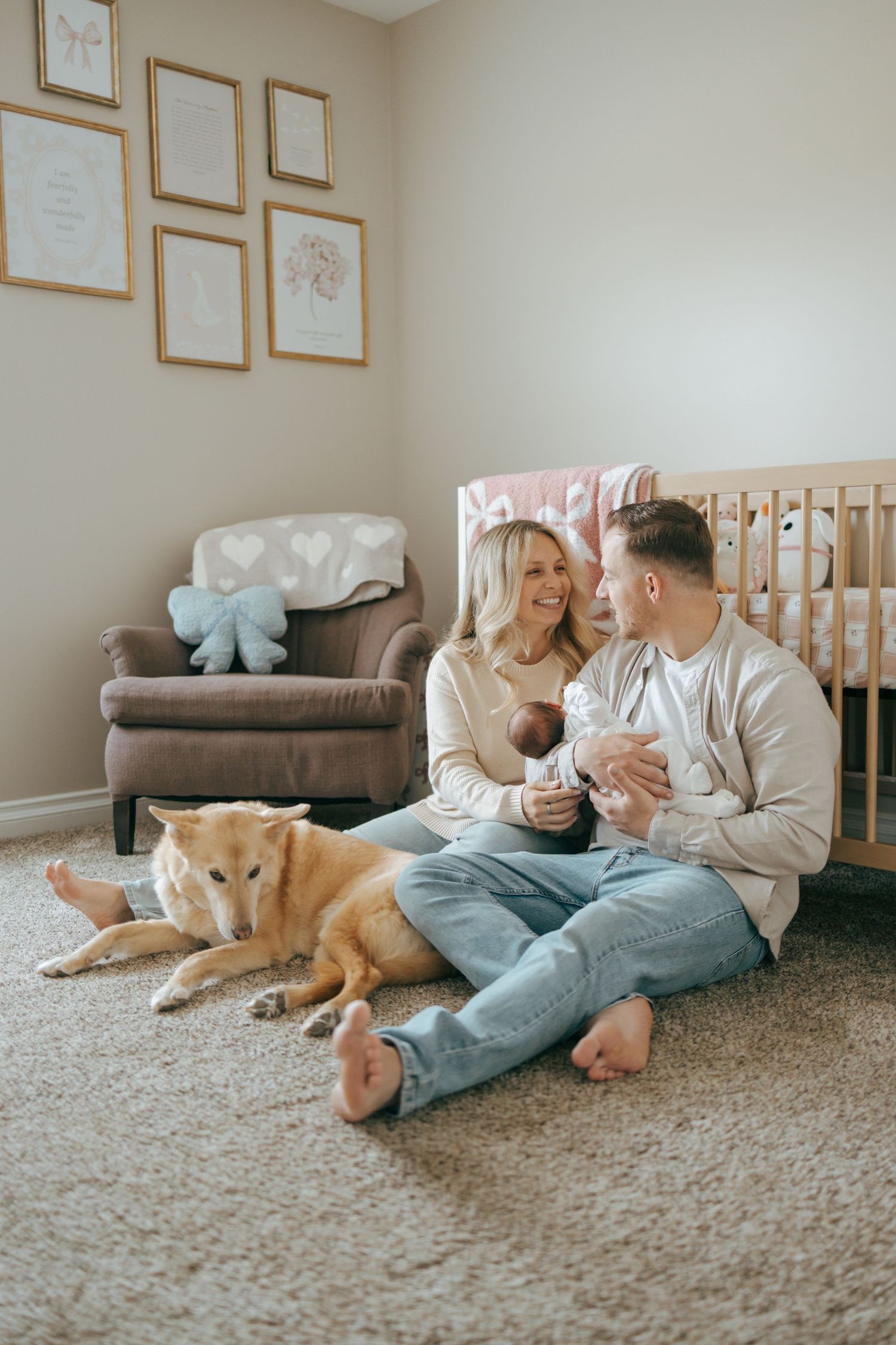 Young family with newborn baby and golden dog sitting on nursery floor by wooden crib.