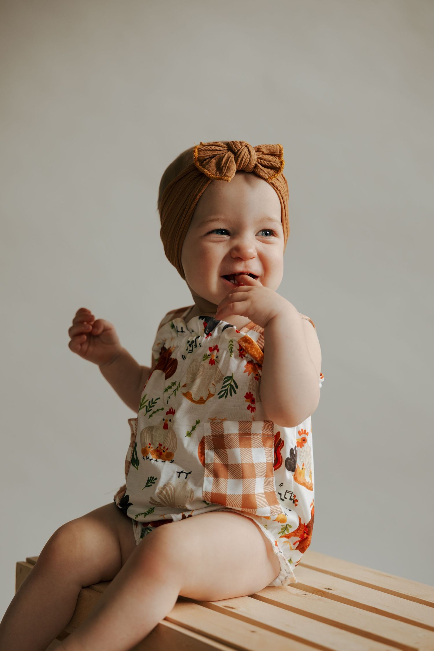 Adorable baby girl in floral romper and mustard bow headband sitting on wooden crate.
