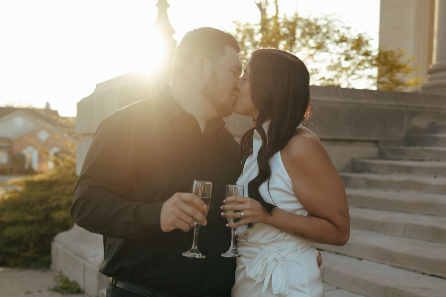 Newlywed couple sharing a kiss while holding champagne flutes in golden sunset light on steps.