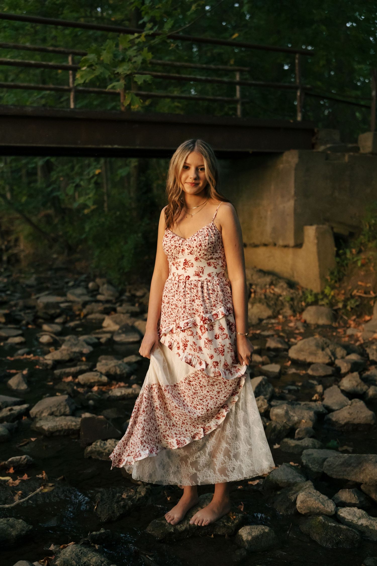 Young woman in white floral maxi dress standing barefoot on rocks beneath a bridge.