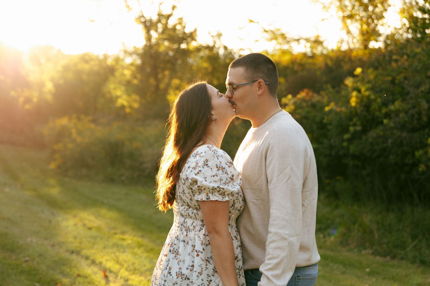 Couple shares a kiss at golden hour sunset in a lush green outdoor setting.