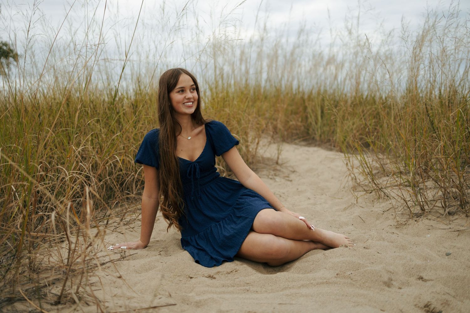 Smiling young woman in navy blue dress sitting on sandy beach path surrounded by tall grass.